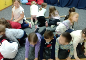 Liczna grupa dzieci siedzi na podłodze w prostokątnej konstrukcji z tekturowych, długich tub.
